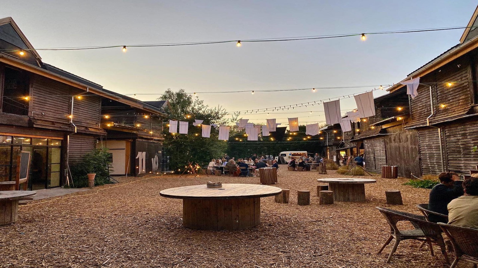Banegaarden barn courtyard at dusk with string lights, white flag bunting, and guests dining at long tables