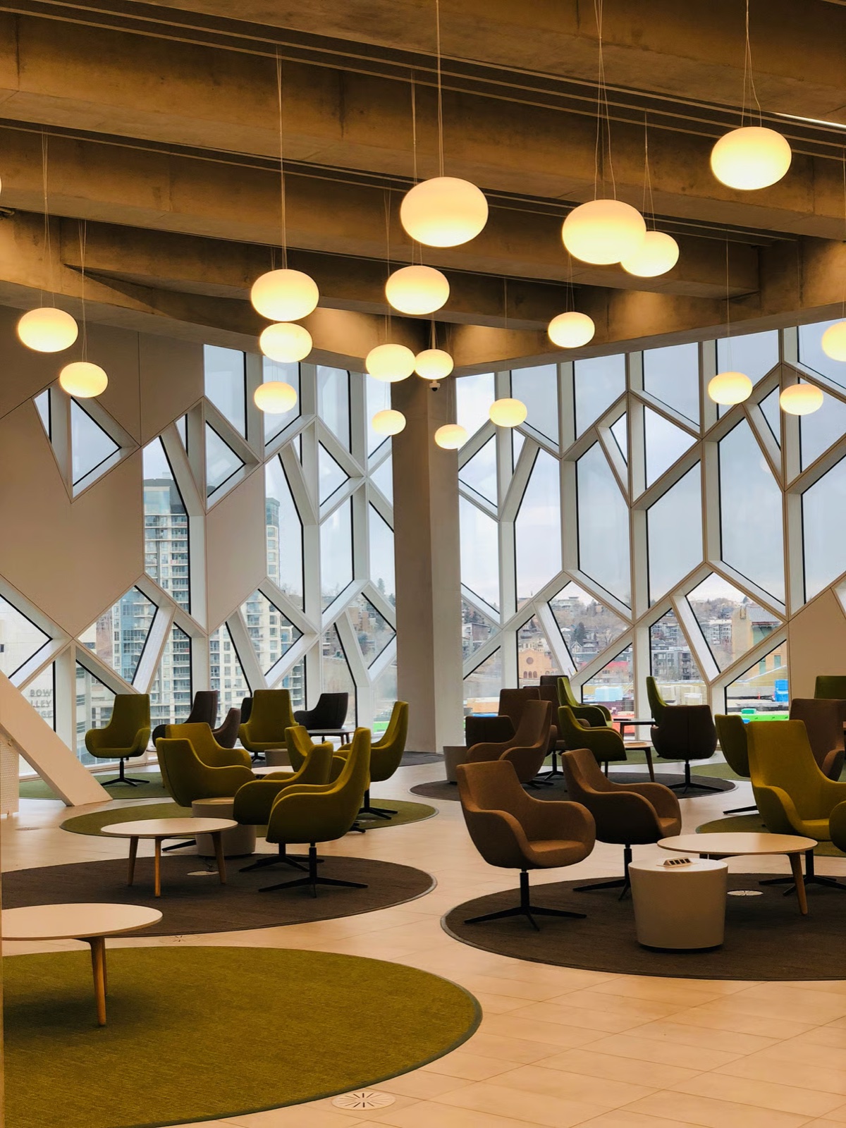 Modern lounge with green and brown swivel chairs, globe pendant lights, and geometric windows overlooking a cityscape